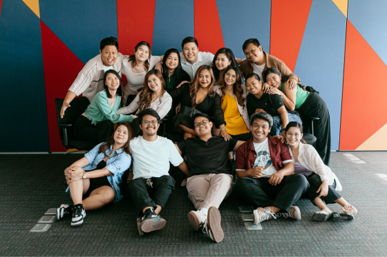 A diverse group of about 18 young professionals, both men and women, are posing together in a cheerful and friendly manner. They are arranged in two rows with some seated on the floor and others standing or leaning behind them. The background features a colorful geometric wall with red, blue, yellow, and orange shapes. Everyone is smiling and appears to be enjoying the moment, showcasing a vibrant and inclusive team atmosphere.