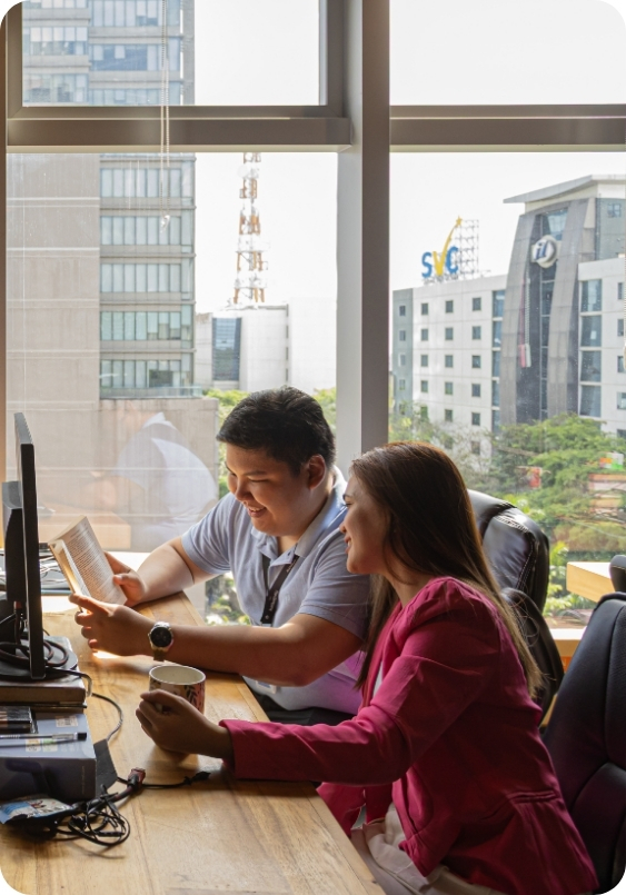 Makati City Virtual Office The Company Makati (1) A cheerful pair works side by side at a wooden table, coffee mugs in hand, with high-rise buildings visible through large windows at The Company Makati. This scene highlights the comfort and credibility of using a Virtual Office in Makati.