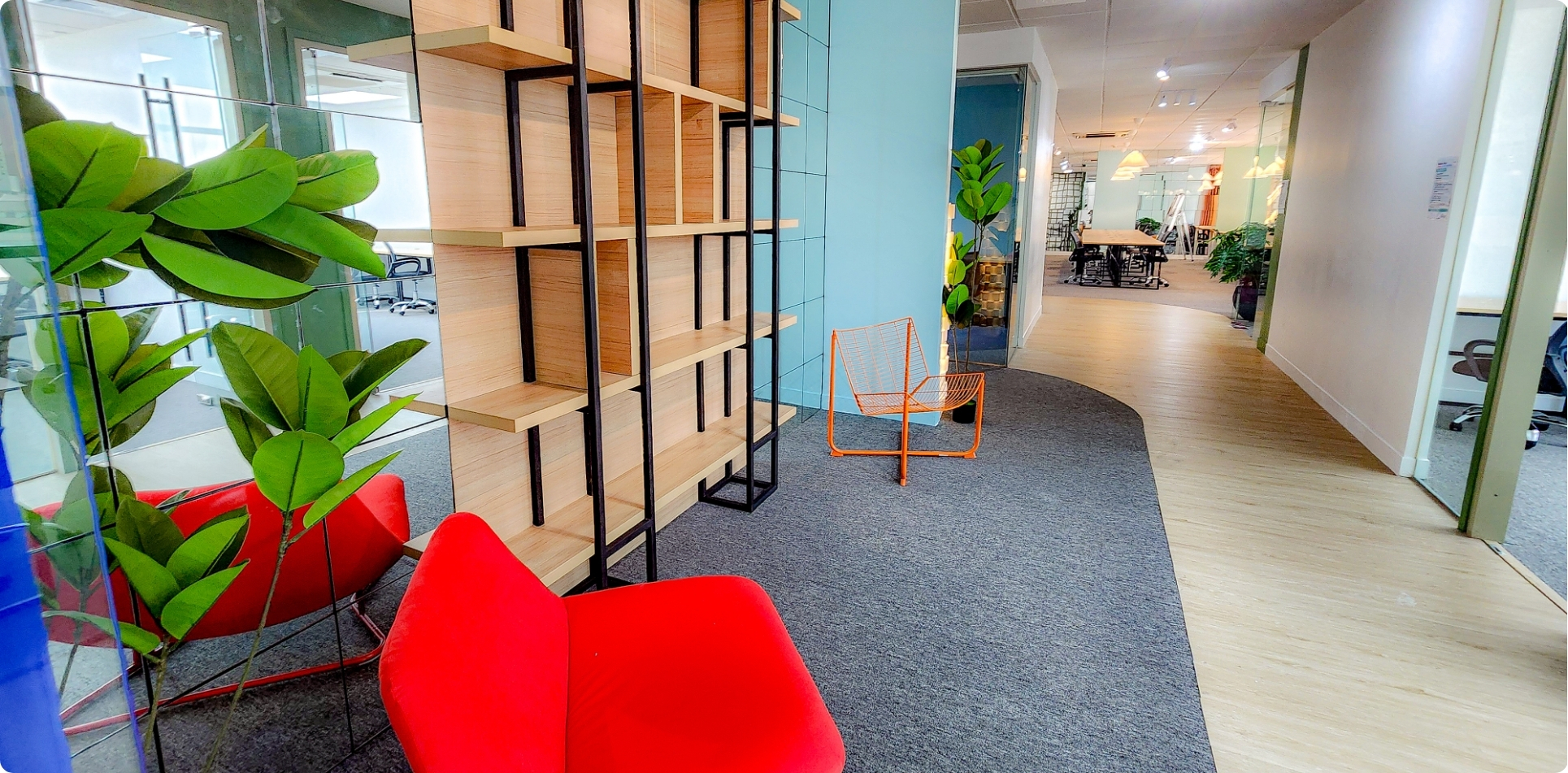 A stylish lounge area at The Company Makati featuring modern red and orange chairs, a wooden open shelf, indoor plants, and a clean, well-lit workspace corridor. This inviting setting highlights the Virtual Office in Makati, perfect for professionals seeking flexible business solutions.