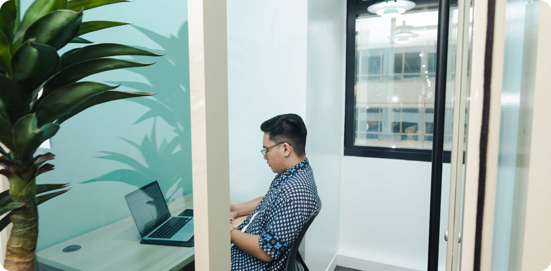 A man wearing glasses and a patterned shirt works on a laptop inside a small, brightly lit private office pod or focus room. To the left, a large, leafy plant provides a biophilic element against a pale turquoise wall. This design, emphasizing quiet focus zones and wellness elements like plants, represents the evolving Future of Commercial Real Estate towards experiential and neuro-inclusive workspaces.